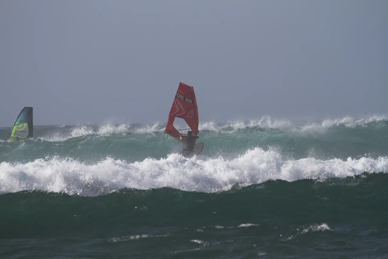 Windsurfer met rood zeil voor een witte golf